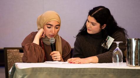 Two people sit at a table. One person reads from a paper, holding a microphone up to their mouth. The other watches on and holds the paper for them