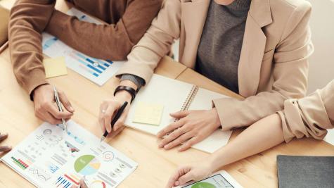 A group of people at a table. They are surrounding charts and hold pens to mark up their papers.