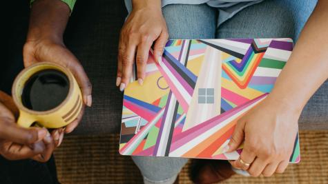 A laptop with a colourful Pride cover laying on someone's lap. A mug of coffee held in the hands of another.