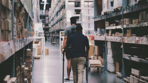 Two men maneuver a trolley in a large warehouse filled with boxes and shelves.