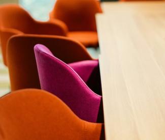 A series of chairs at a table. Orange chairs surround one singular pink chair.
