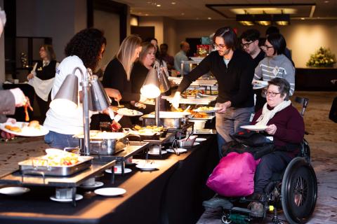 A group of people at a buffet serving themselves. A wheelchair user holds a plate while another person serves them food