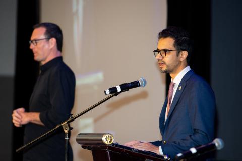 Two speakers stand onstage. One is in front of a podium speaking into a microphone while their image is projected behind them onscreen 