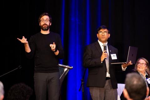 One speaker stands onstage holding a microphone and a laptop. A sign language interpreter stands beside them signing to the audience