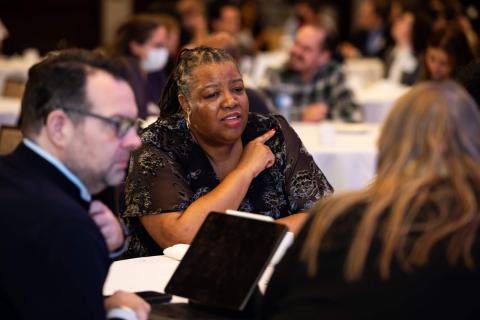 A table of people talk to one another. One person is gesturing off camera while another sits in front of a tablet