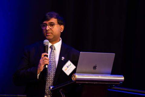 A person stands onstage at a podium. They hold a microphone and have a laptop resting on the podium in front of them