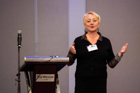 A person wearing a nametag stands beside a podium holding their hands up as they speak to the audience