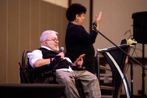 A person in a motorized wheelchair holds a microphone and a phone in his hands, speaking to the audience while a sign language interpreter stands to their left at a podium and signs to the audience