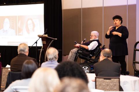 An audience view of the stage. The audience listens to a person in a motorized wheelchair speak while a sign language interpreter signs what they say to the audience