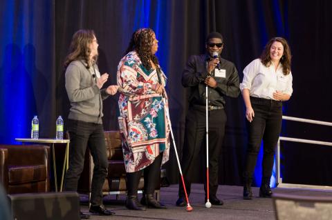 Four people stand onstage. Two people hold folding canes while one of them speaks into a microphone. 