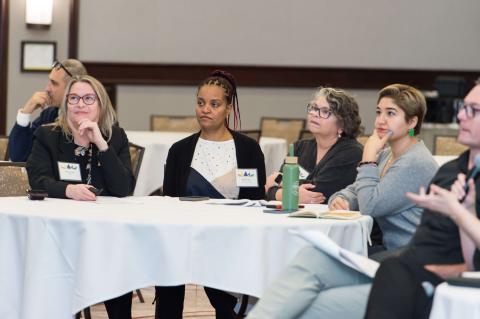 Four people sit a table staring ahead at a speaker off-camera.