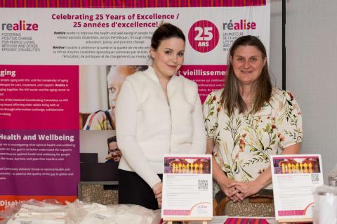 Two people stand at an information table. A large white, pink, and purple display hangs behind them.