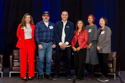 A group of six standing onstage in front of a blue curtain. They smile and pose for photos.