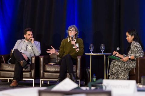 Three speakers sit onstage. One of them speaks into a microphone while the other two listens.