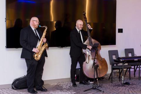 Two band members play an upright bass and a saxophone in front of a large screen