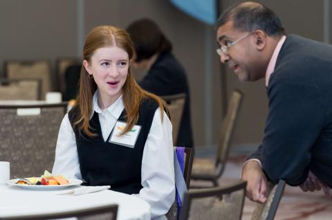 Two people conversing at a table. One person leans on a chair while the other sits by their plate of food.