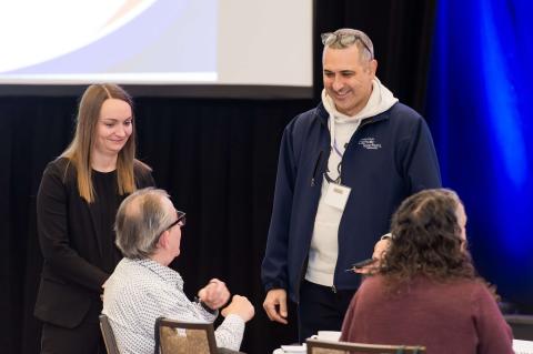 Four people talk around a table. Two stand in front of a stage with a blue curtain while the other two sit at a table with a white table cloth.