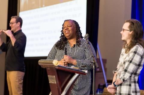 Two speakers standing on stage in front of a screen and a blue curtain. A sign language interpreter translates for the audience close by.