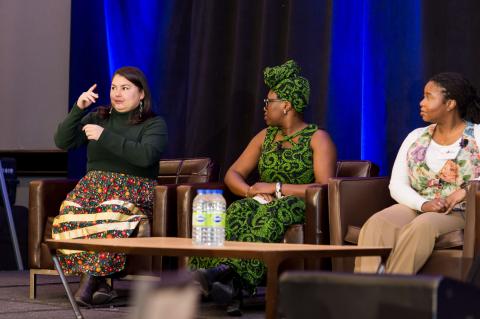 Three speakers sit on stage in front of a blue curtain. Two of them watch the other speaker as they address the audience.