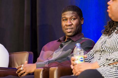 A close up portrait of two speakers sitting on stage. One of them watches the other speak.