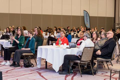 An audience sitting at tables with white tablecloths. They sit in a room with a patterned carpet and brown and tan walls.