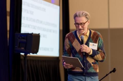 A speaker standing on stage holding a microphone and reading from a tablet.