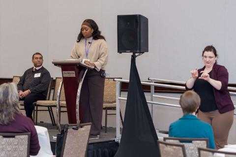 A speaker standing at a podium with another speaker sitting on a chair to their right. To their left, an interpreter signs to the audience. 
