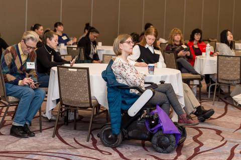 An audience sits at tables in chairs and wheelchairs, listening to the speakers.