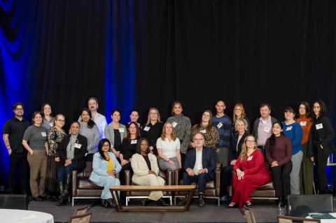 Twenty-five speakers pose on stage in front of a blue curtain. They are all arranged for the photo, some sitting and some standing behind.