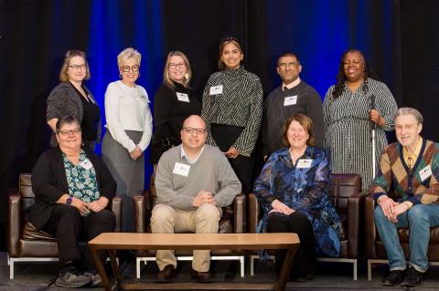 Ten speakers pose for a picture on stage. Four of them sit in brown chairs while the other six stand behind them