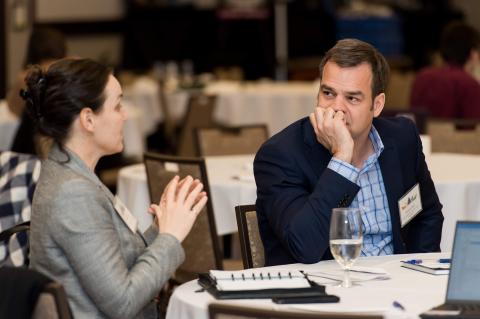 Two people sit and talk at a table with notebooks in front of them.