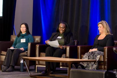 Three speakers sit on stage in brown chairs and in front of a blue curtain.