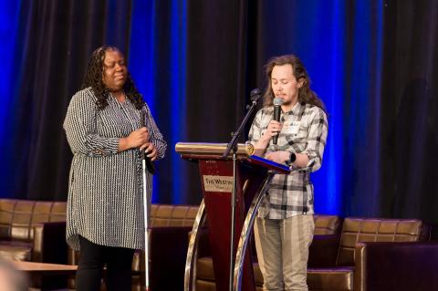 Two speakers stand on stage at a podium. Both hold microphones and one watches the other speak.