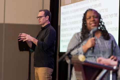 A speaker addresses the audience with a microphone. Nearby, a sign language interpreter signs to the audience.