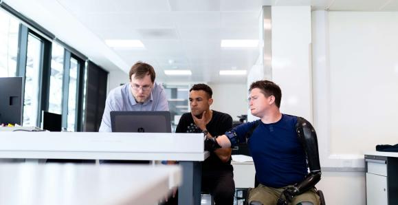 colleagues working at a computer; on is standing, one is seated in a chair and one is seated in a wheelchair 