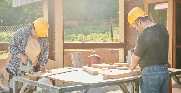 Two construction workers working at a saw bench. They are wearing hard hats.