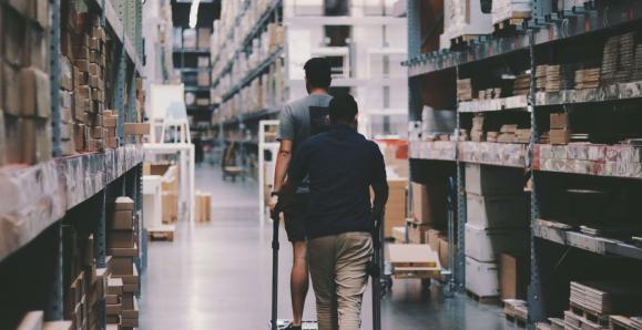 Two men maneuver a trolley in a large warehouse filled with boxes and shelves.