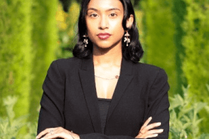 Portrait of Ronessa Dass standing outdoors against a green leafy background, wearing a black outfit with arms crossed and looking at the camera.