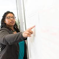 a black woman stands at a whiteboard giving a lesson