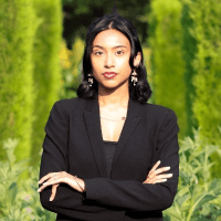 Portrait of Ronessa Dass standing outdoors against a green leafy background, wearing a black outfit with arms crossed and looking at the camera.