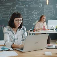 A classroom with a chalk board in the background. In the foreground, People work at laptops at desks. 
