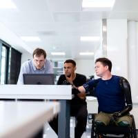 colleagues working at a computer; on is standing, one is seated in a chair and one is seated in a wheelchair 