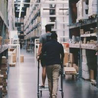 Two men maneuver a trolley in a large warehouse filled with boxes and shelves.